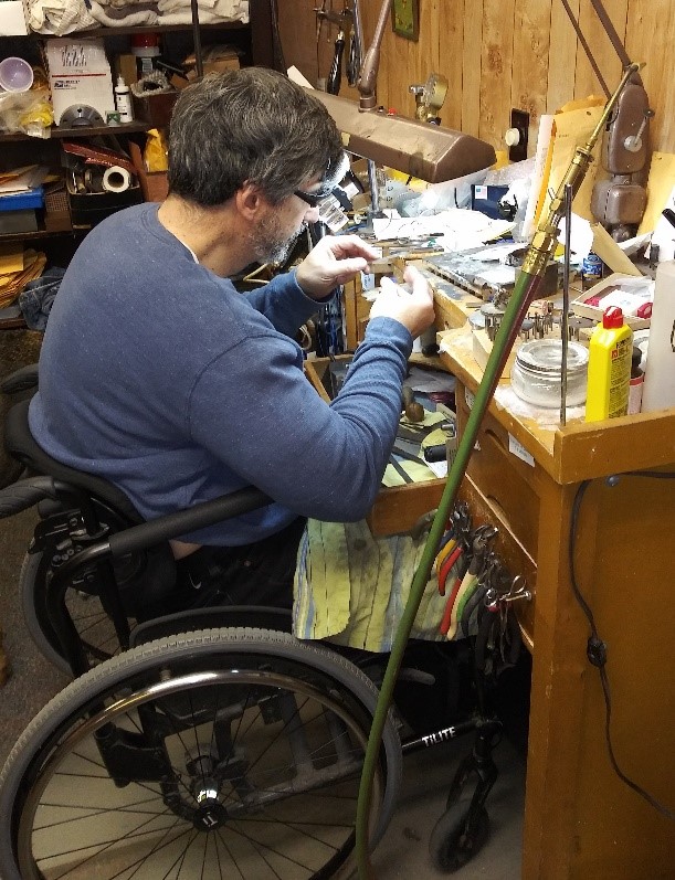 Photo of Dan Banda sitting in his manual wheelchair working on a piece of jewelry at his workbench.