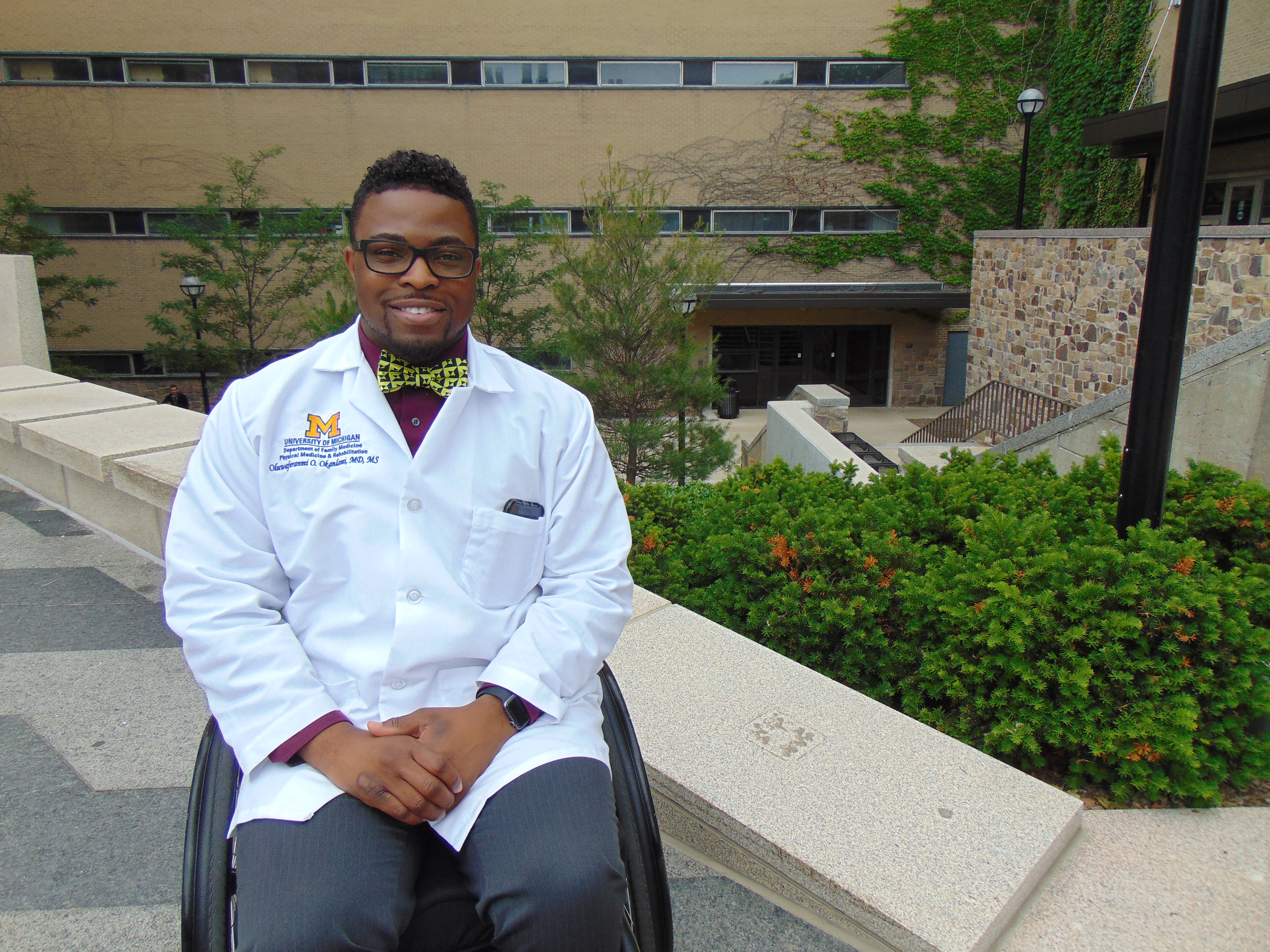 Dr. Oluwaferanmi Okanlami poses in his wheel chair in front of the U-M Medical School. He is wearing a white physician's coat. 