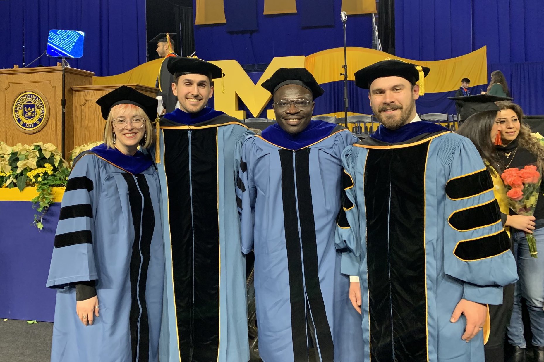 Four graduate students in cap and gown, at graduation ceremony