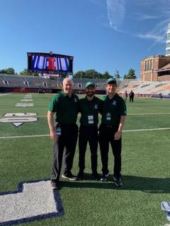 Three male physicians standing in the middle of a football field. They are each wearing dark green polo shirts.