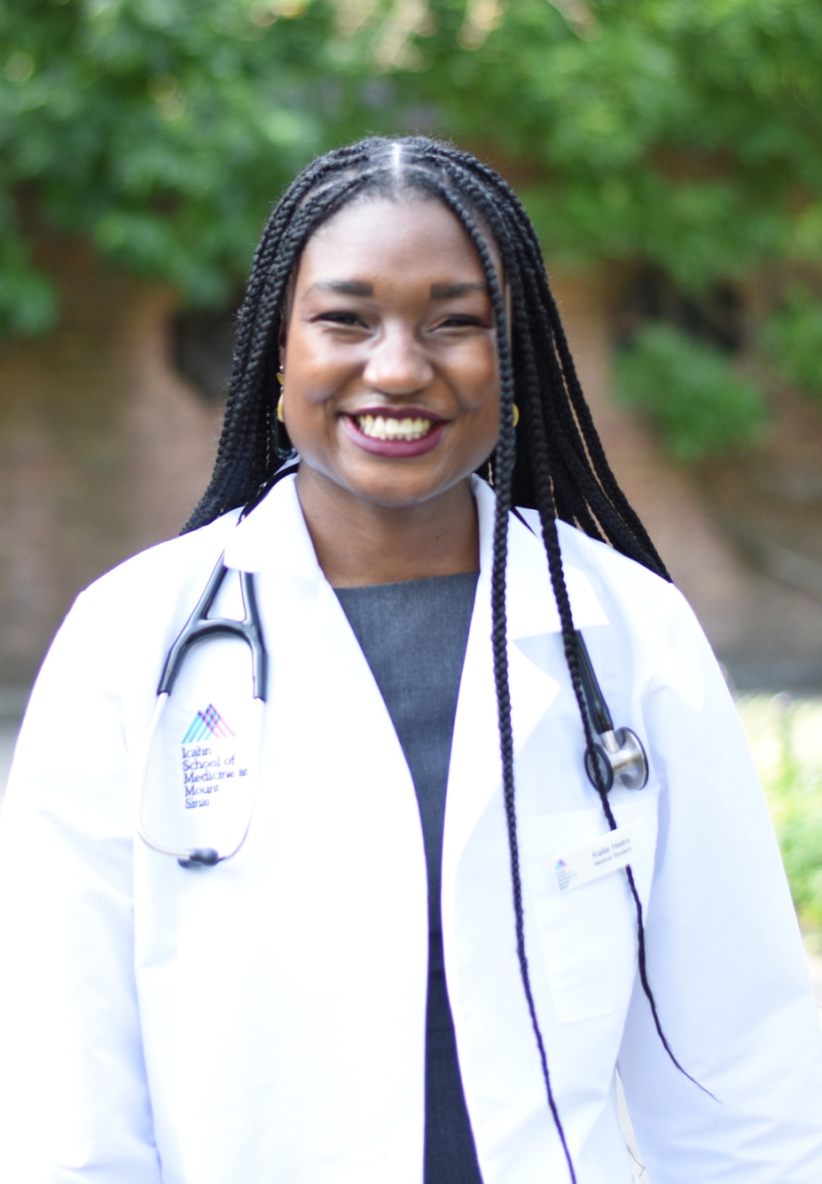 A photo of Kaila Helm wearing a white medical coat and standing in a green garden.