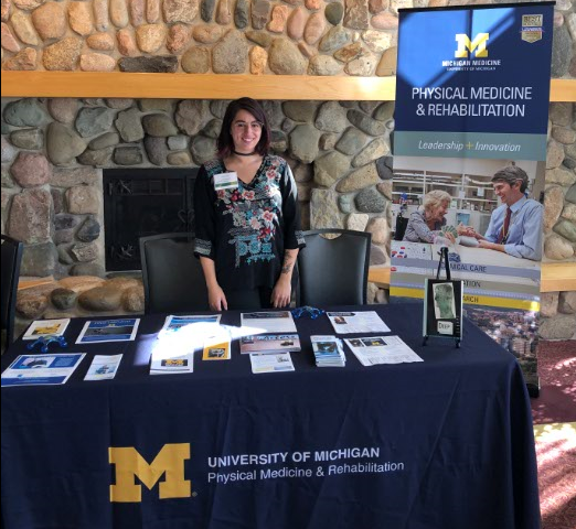 Photo of Lynn Charara, one of our research associates standing at a research exhibition table at the Mary Free Bed Spinal Cord Injury Symposium in Grand Rapids on September 26, 2019.
