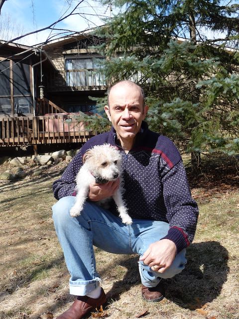 Philip Zazove kneeling with one knee on the ground, holding a small white dog on his other knee. He is outside and smiling at the camera.