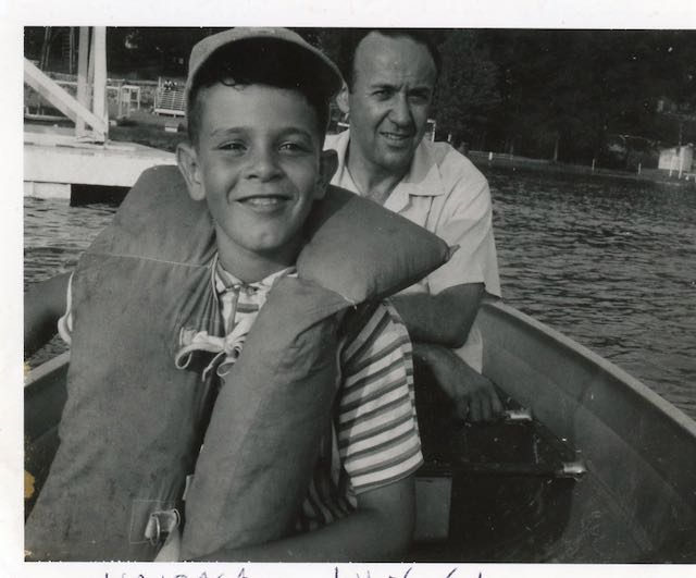 Black and white photo of a young boy wearing a life jacket, a man peeks from behind him. They are in a boat. 