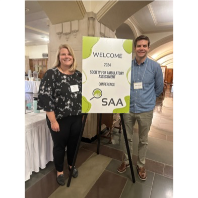 Two people standing next to a sign that says welcome to the 2024 society for ambulatory assessment conference