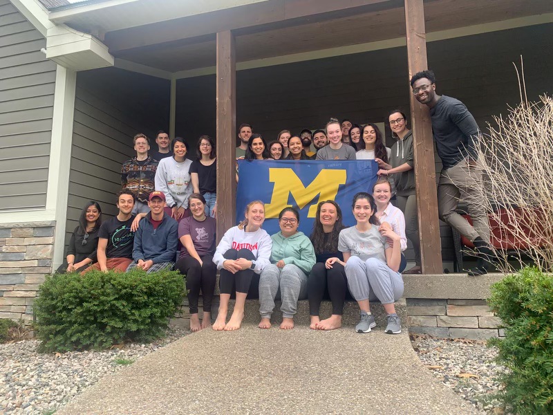 A group of resident physicians standing on the porch of a tan colored house. They are holding a U-M flag that is blue with a maize M on it. 
