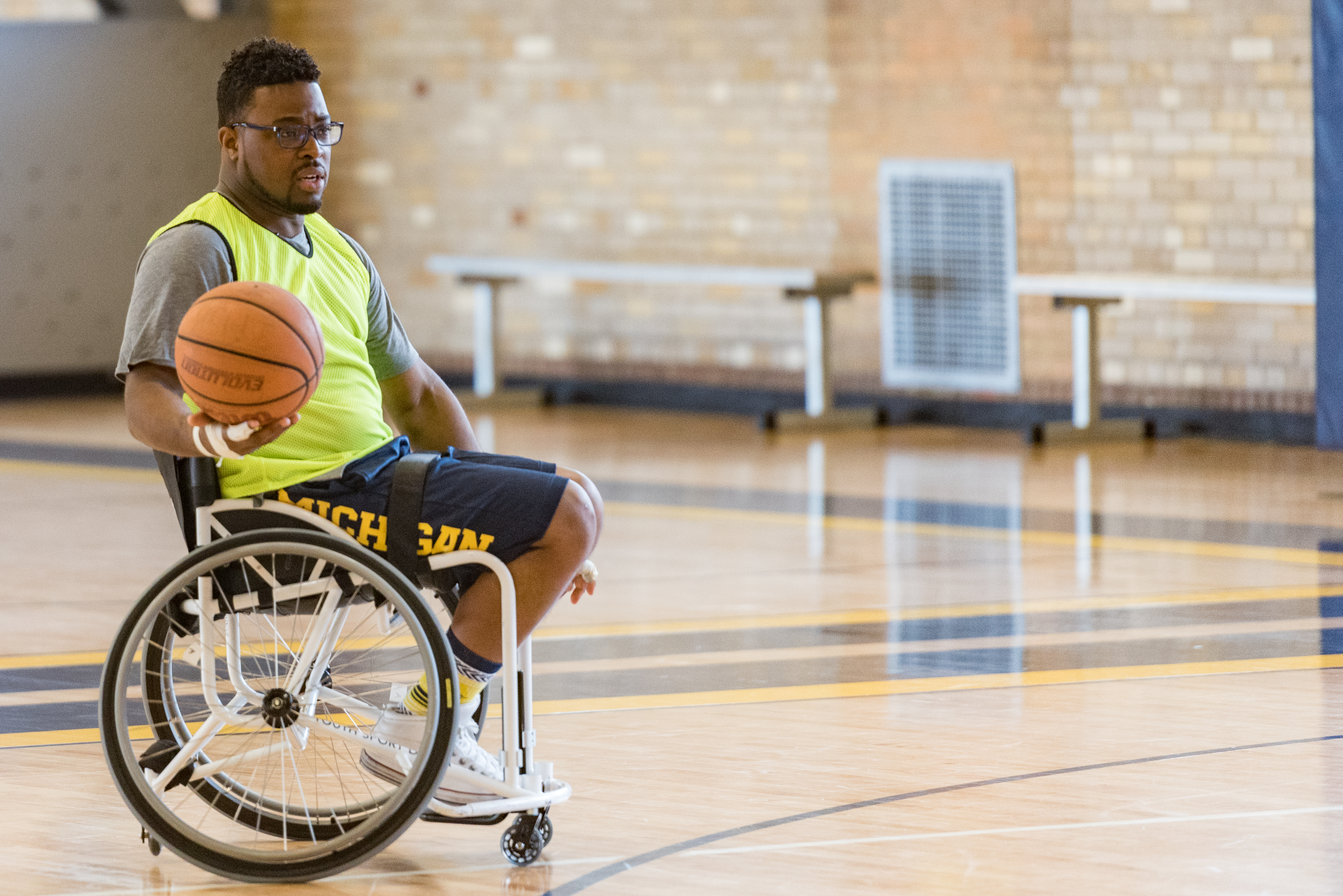 Dr. Feranmi Oklanmi sits in a sports wheel chair and holds a basketball in one hand. He is on a basketball court. 