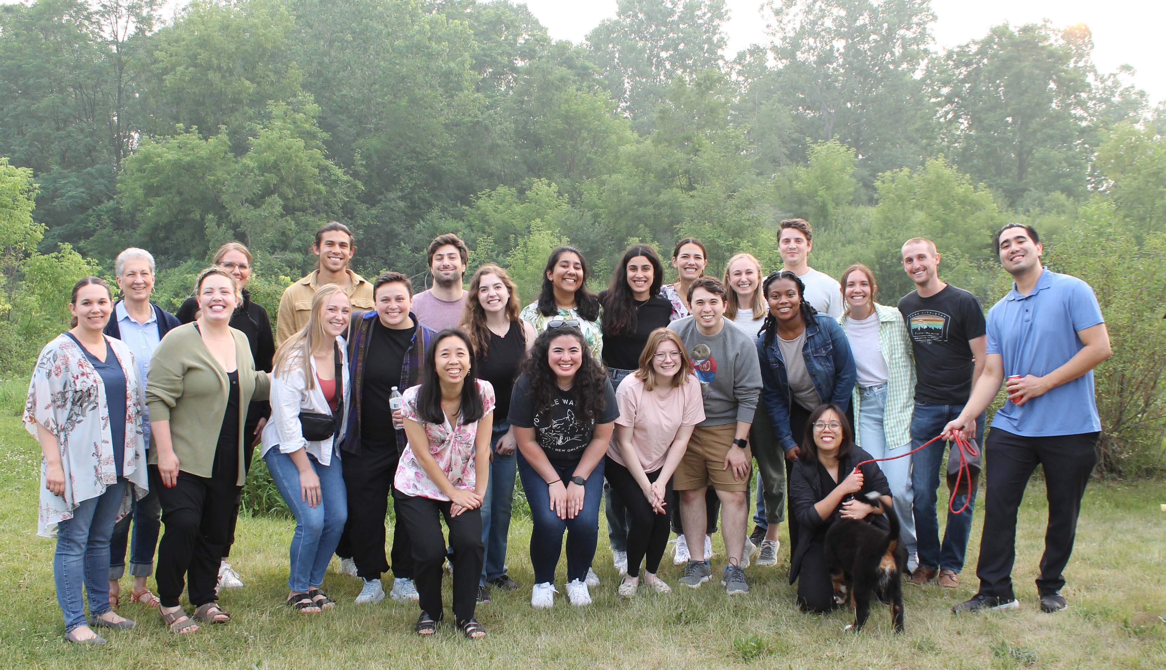 A group of 23 people stand gathererd together looking at the camera and smiling. They are standing outside in front of green trees. One holds a dog leash with a black dog. 