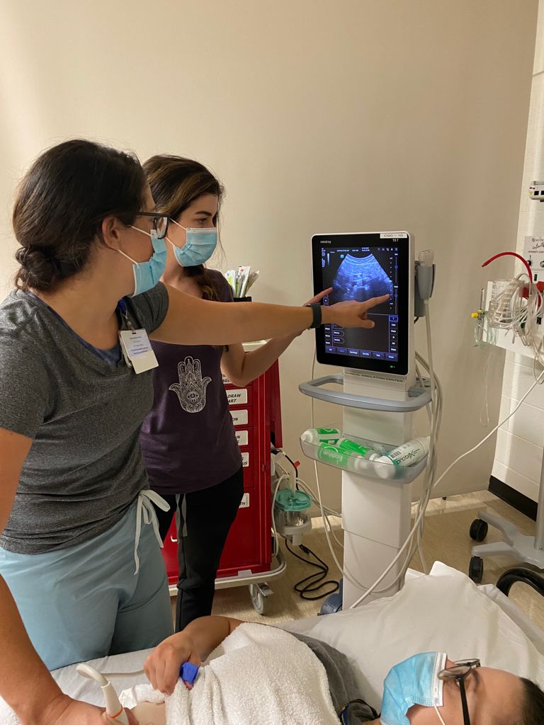 Two female resident physicians in masks point at an ultrasound screen. One resident physician lies on a hospital bed and also looks at the screen