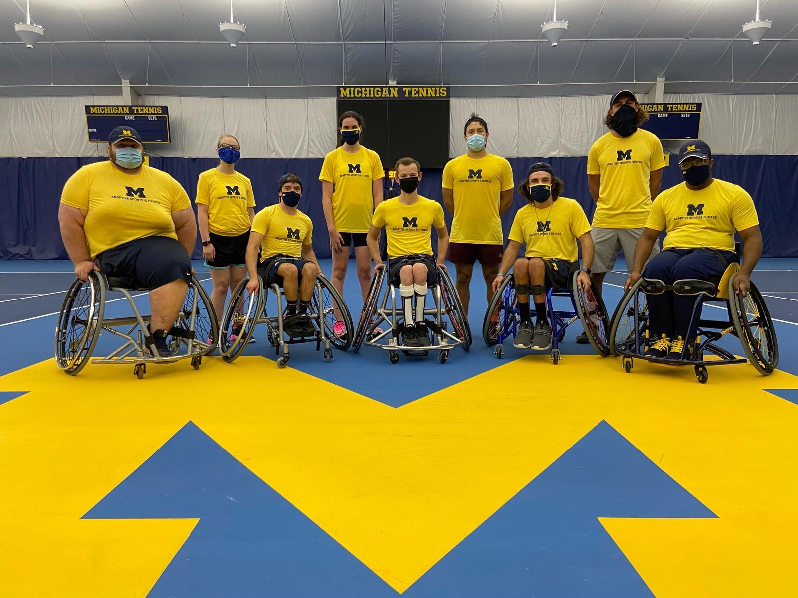 ASAF Tennis Team - Nine athletes in two rows in masks and yellow t-shirts with Block M's. The athletes in the front row are in wheelchairs and the back row is standing. They are on an indoor U-M tennis court. A sign behind them reads "Michigan Tennis"