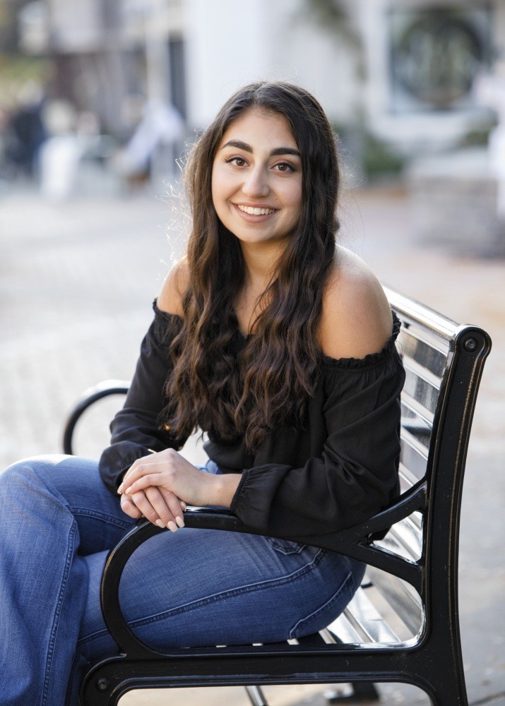White woman, black hair, smiling, wearing a black off the shoulder top, blue jeans, sitting on a bench outside.