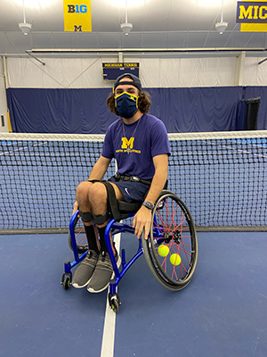 Man wearing a fabric mask in an athletic wheelchair on a tennis court in front of a tennis net. 