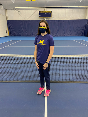 Woman wearing a fabric mask stands in front of a tennis net on a tennis court. 