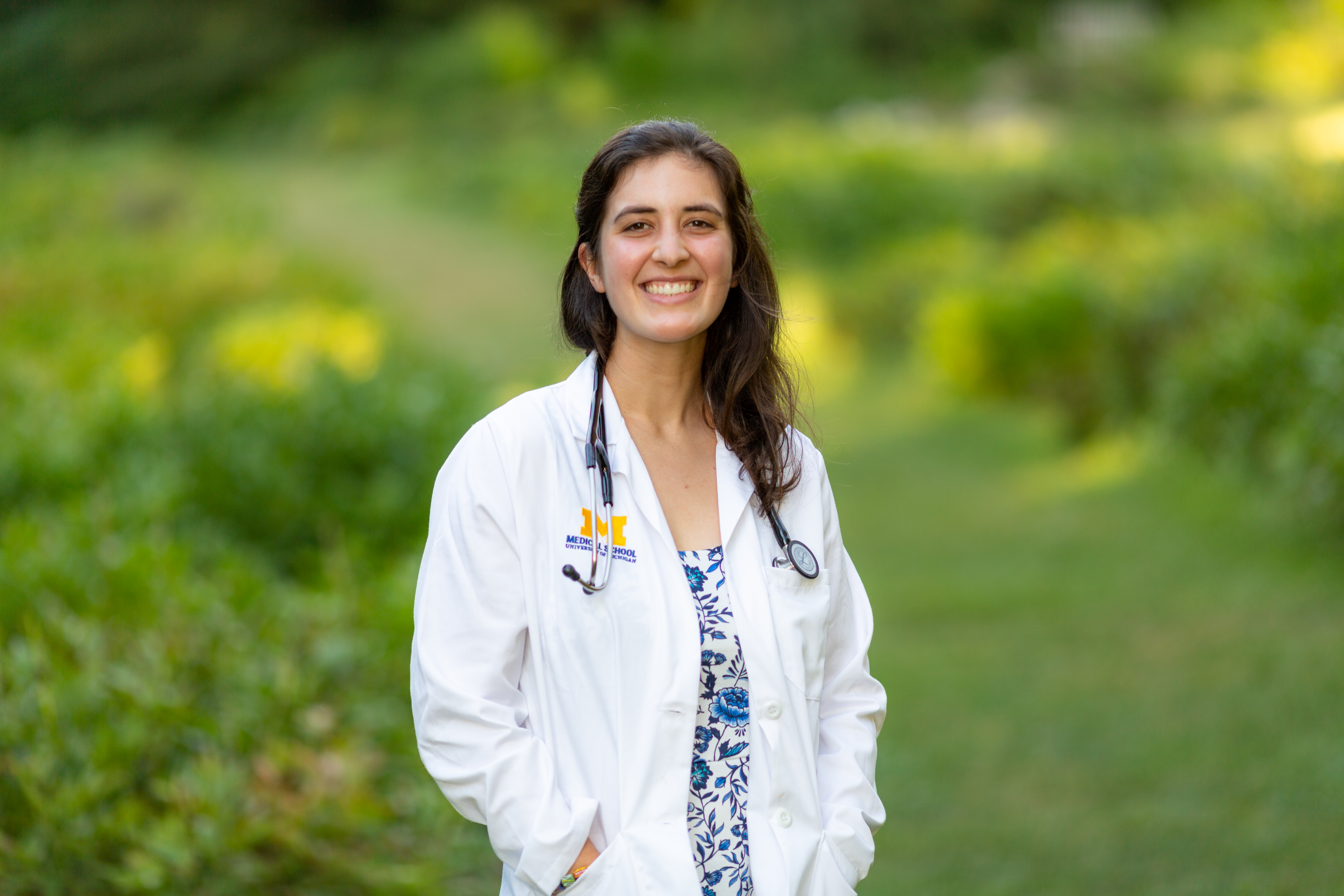 Kate is a white female with dark hair, she is wearing a blue flower print dress, white lab coat and a stethoscope around her neck. The background is a blurred green landscape.