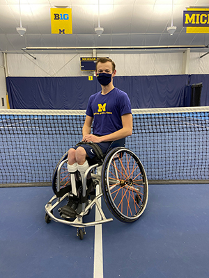 Man wearing a fabric mask in an athletic wheelchair on a tennis court in front of a tennis net. 