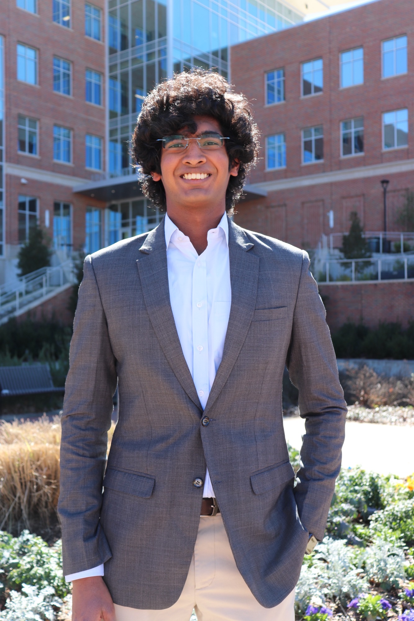 Indian male with brown curly hair, with glasses, smiling and wearing a gray suit coat over a white shirt with khaki pants standing outside with a building behind him.