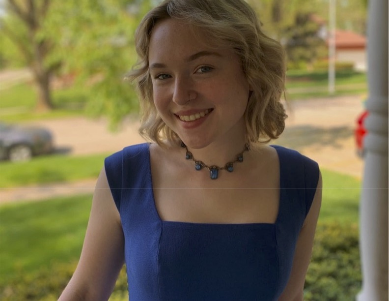 White female, with short blonde hair, smiling, wearing a blue shirt with a necklace. She is outside, blurred background.