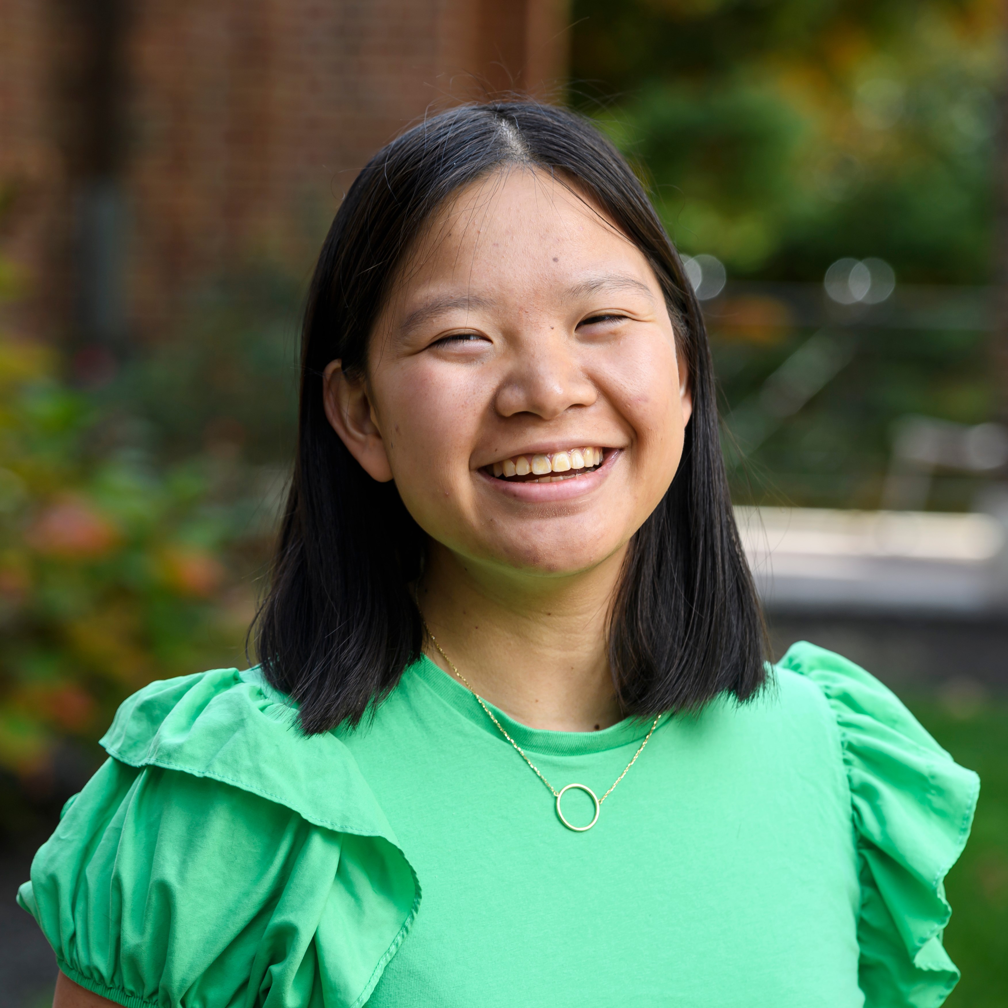 Female, Asian descent, with black short hair, smiling, wearing a green short sleeved shirt, necklace, outside blurred background.