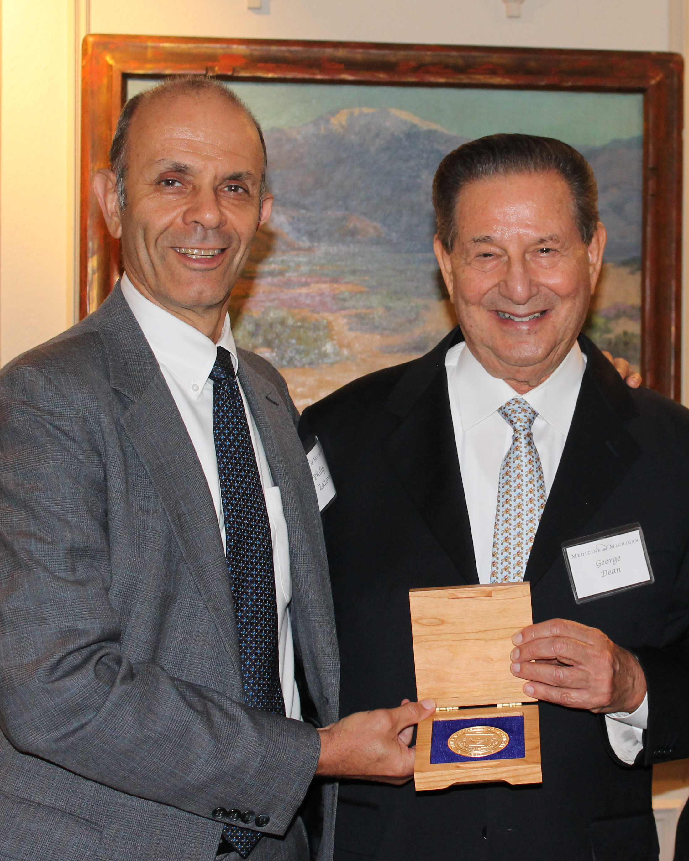 Two men in suits hold a gold medallion in a box between them. They are smiling at the camera. 