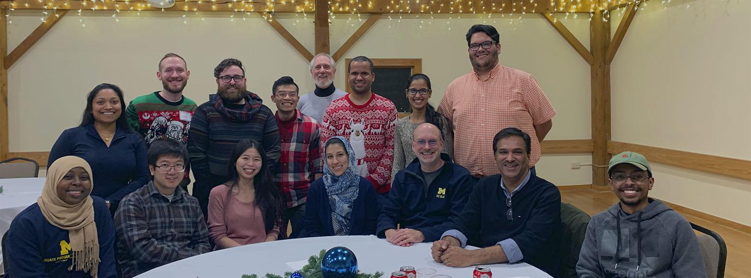 Group shot of 15 smiling adults seated around a table, some wearing holiday sweaters with twinkle lights in the background