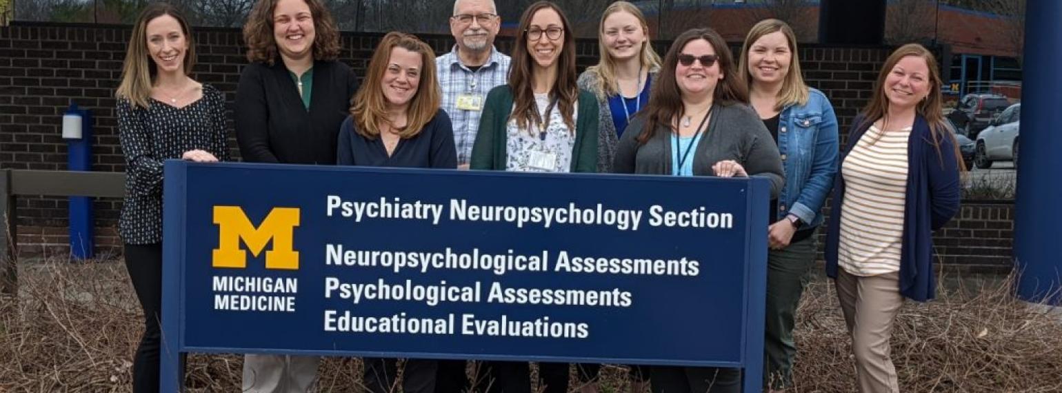 A group of Michigan Medicine employees are standing around a building sign that says "Psychiatry Neuropsychology Section and Neurospychological Assessments, Psychological Assessments, and Educational Evaluations"