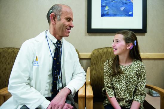 Dr. Philip Zazove sits next to a young girl. They have their heads turned toward each other and each of their cochlear implants are visible 