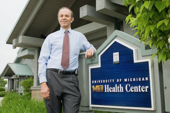 Philip Zazove standing outside, in front of a building, wearing a dress shirt and tie, leaning on a sign that reads University of Michigan Dexter Health Center