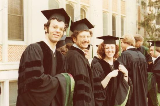 Two men and one woman in black doctoral caps and gowns smile at the camera. They are outside and the picture is an old snapshot taken in 1976.