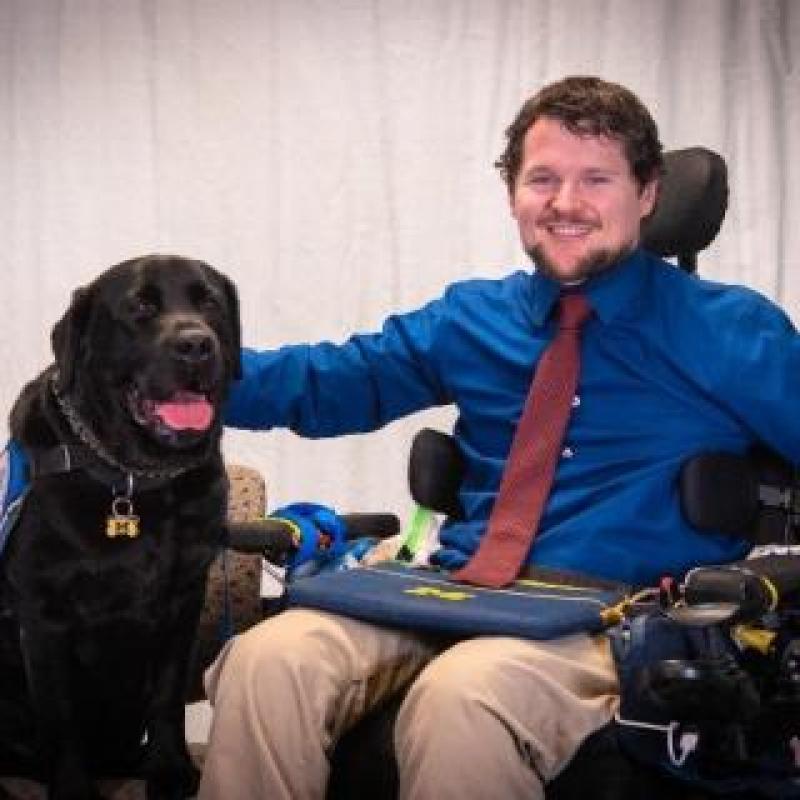 Photo of James Murhta, an individual with a SCI. He is sitting in his power wheelchair with his service dog at his side.