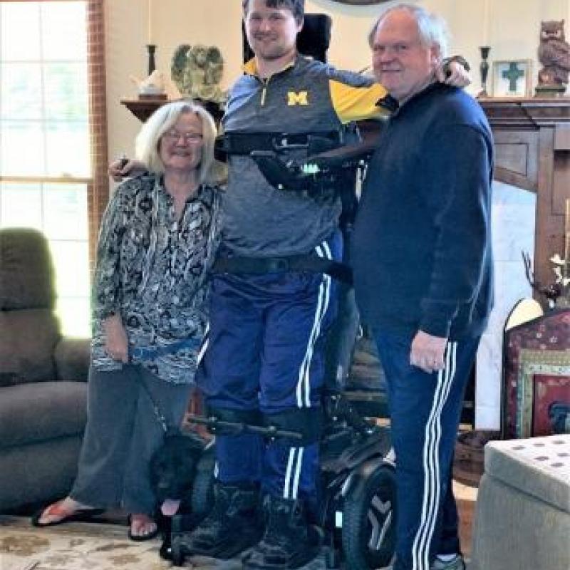 Photo of James with his parents.He is standing upright in his power standing wheelchair.