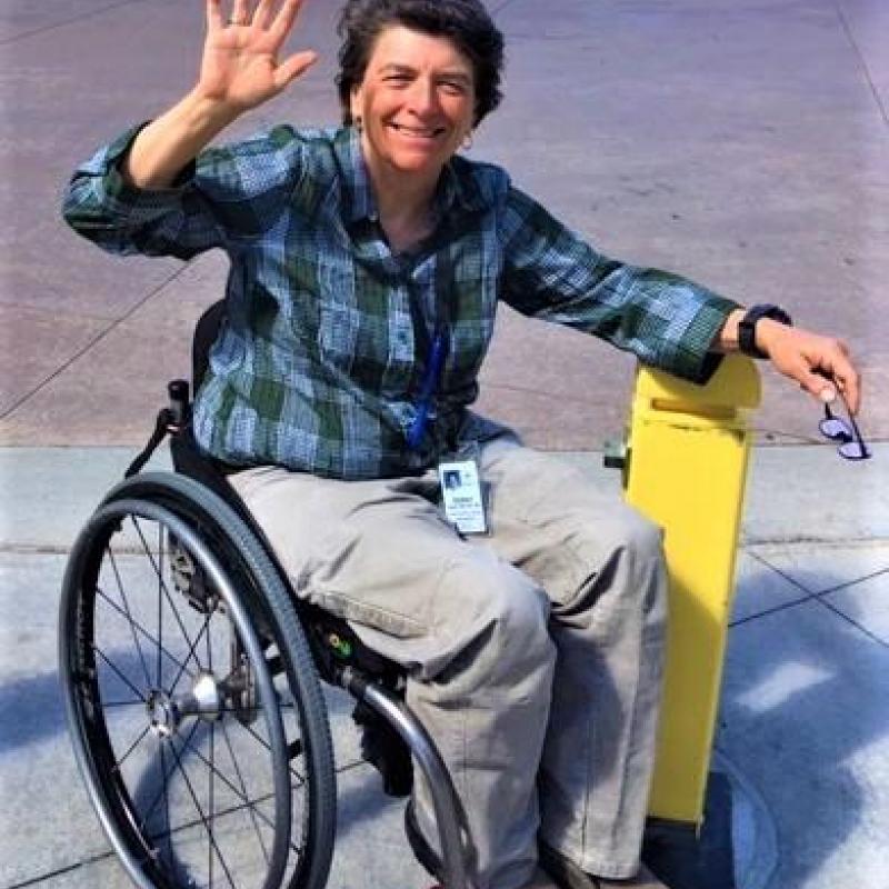 Photograph of Terry Chase, an individual with a spinal cord injury. He has short dark hair and is smiling and waving towards the camera while sitting in a manual wheelchair.