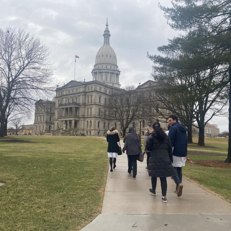 A group of people walk on a sidewalk away from the camera towards Michigan's capitol building 