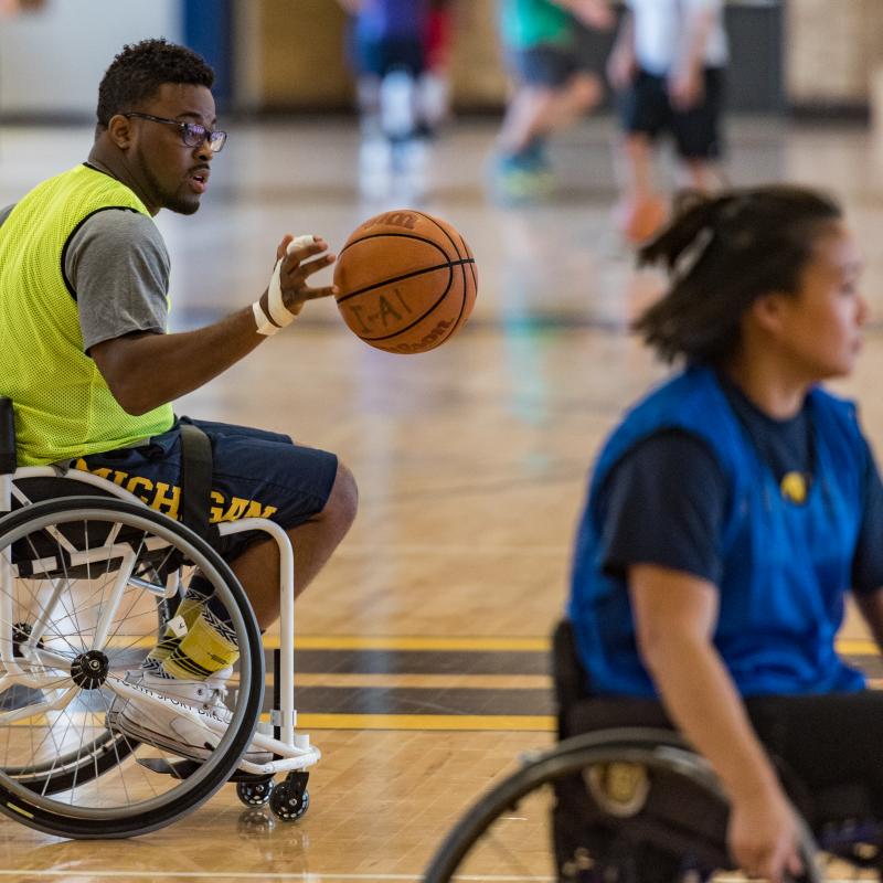 Dr. Okanlami dribbles a basketball as he rolls across a basketball court behind a woman also in a sport wheelchair. 