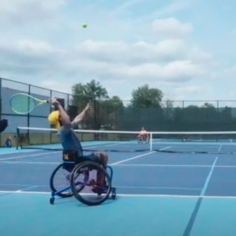 An adaptive tennis player with his back to the camera raises his tennis racket to hit a flying tennis ball