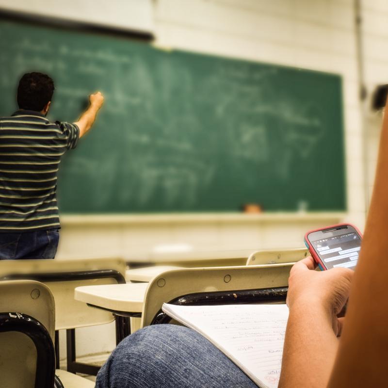 In the background a man stands at a green chalkboard. He is writing on the board and his back is to the camera. In he foreground a close up of a person at a desk. You see the side of their arms and lap and they are holding a smartphone that is turned on. 