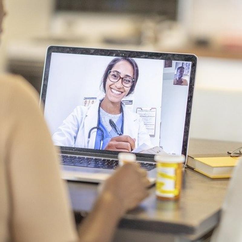 woman writing notes while talking to her doctor via laptop, doctor is in white coat and smiling with glasses, woman has her prescription medications out
