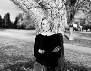 Carey Candrian, Ph.D.  - a black and white photo of a white woman with light hair leaning against a tree