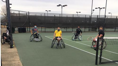 An adaptive tennis team (4 men with athletic wheelchairs) on a tennis court. 