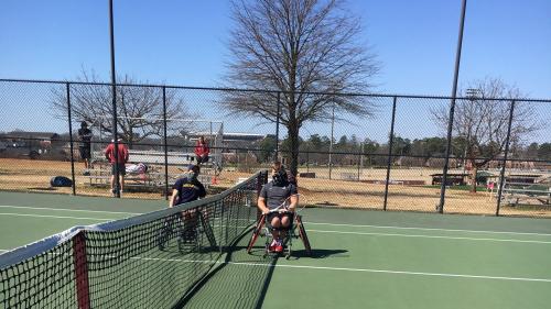 Two men with athletic wheel chairs are on either side of a tennis court. 