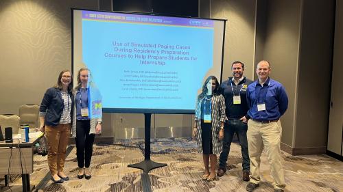 Five people stand in front of a projection screen in a conference room. They are smiling. The screen reads "Use of Simulated Paging Cases During Residency Preparation Courses to Help Prepare Students for Internship."