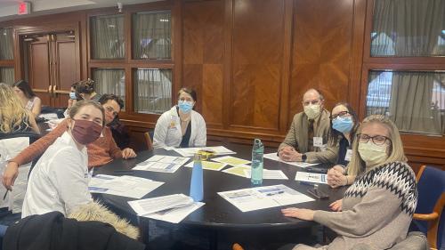 Six people wearing masks sit around around a round table and are turned to look at the camera.