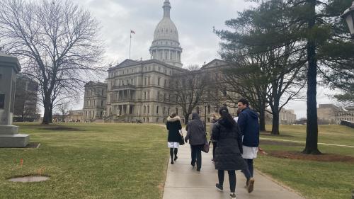 A group of people walk on a sidewalk away from the camera towards Michigan's capitol building 