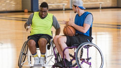 Two basketball players in sport wheelchairs are on a basketball court. One is holding a basketball. 