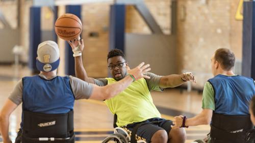 Three basketball players in sport wheelchairs are on a court. One has the ball raised in a hand while another blocks.