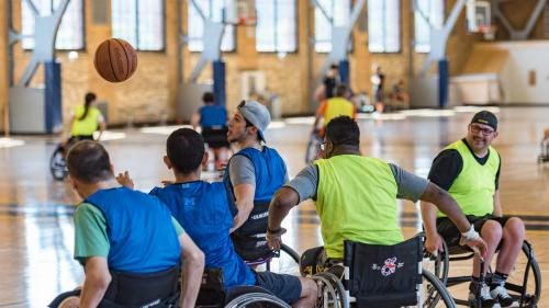 A group of basketball players in sport wheelchairs play a game of basketball on a court. In the background, another group plays on a second court. 